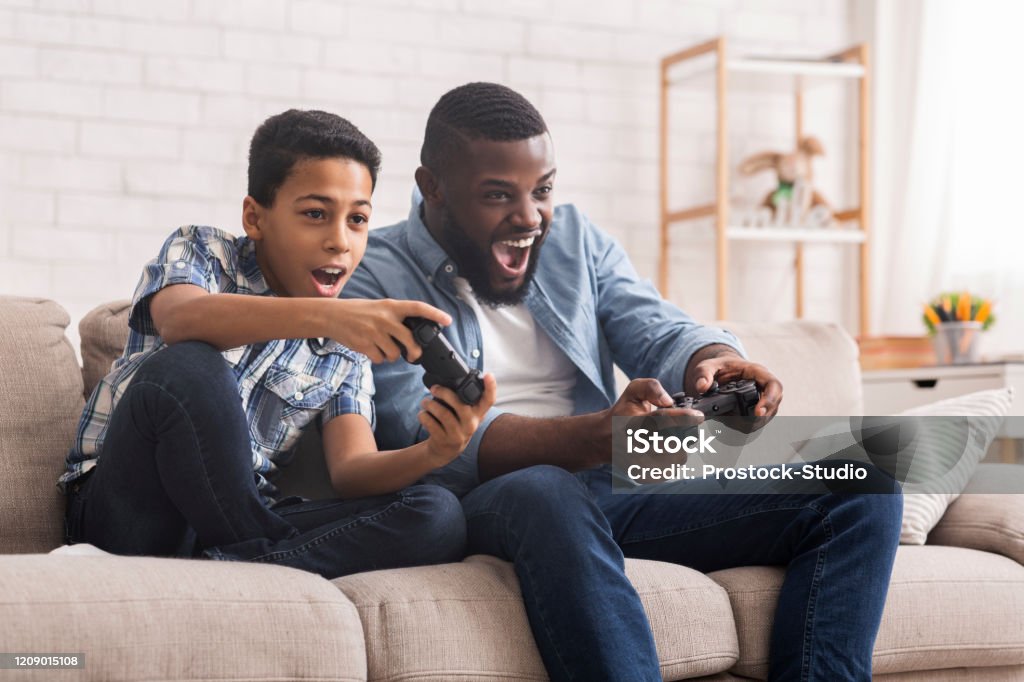 Cheerful Black Father And Son Competing In Video Games At Home Leisure With Dad. Cheerful Black Father And Son Competing With Each Other In Video Games, Using Joysticks, Having Fun At Home, Free Space Video Game Stock Photo