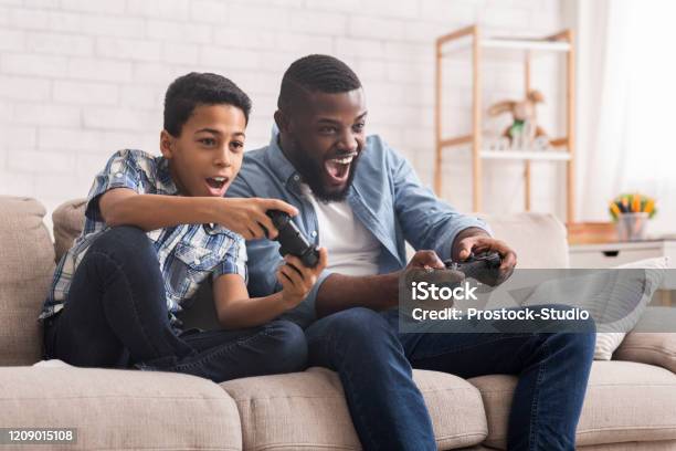 Cheerful Black Father And Son Competing In Video Games At Home Stock Photo - Download Image Now