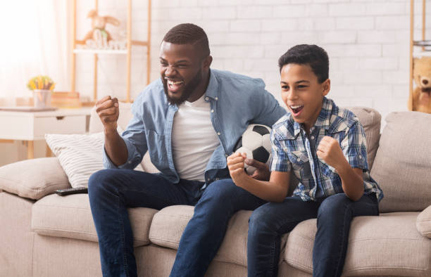 興奮した黒人の父と息子がテレビでスポーツを見て応援する - fan television football watching tv ストックフォトと画像