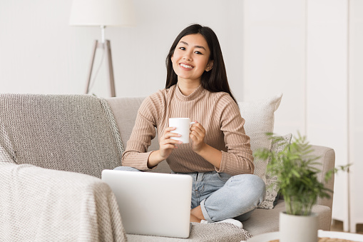 Enjoy weekend. Asian girl relaxing, drinking coffee and using laptop, sittin gon couch at home