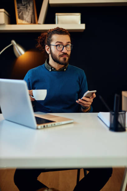 istock Handsome bearded caucasian hipster sitting in his modern home office, using smart phone for checking messages on social media and holding cup of coffee. 1209018340