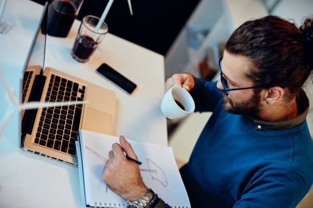 istock Young innovative dedicated bearded employee sitting in his modern office and drawing sketches of windmills and drinking coffee. Sustainable development concept. 1209018518