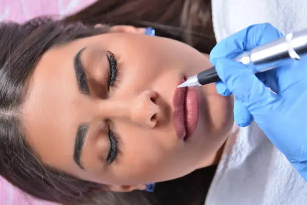 Beautiful and pretty woman having permanent makeup on lips in beautician salon. Beautician hand with tattoo instrument near lips. A girl lies in bed. Close up, selective focus