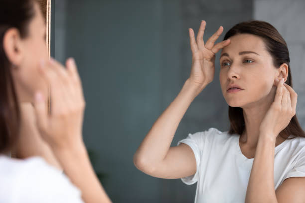 Anxious woman looking in mirror, touching forehead, confused about wrinkles Anxious woman looking in mirror, standing in bathroom, upset attractive female touching forehead with finger, confused about wrinkles or acne, thinking about face lifting procedure or spa forehead photos stock pictures, royalty-free photos & images