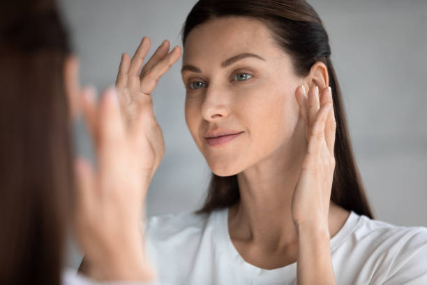 close up beautiful woman with perfect skin looking in mirror - massage creme imagens e fotografias de stock