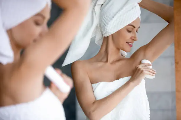 Smiling beautiful woman applying antiperspirant to armpit after shower close up, standing in front of mirror in bathroom, pretty young female wearing white bath towel on head using stick deodorant