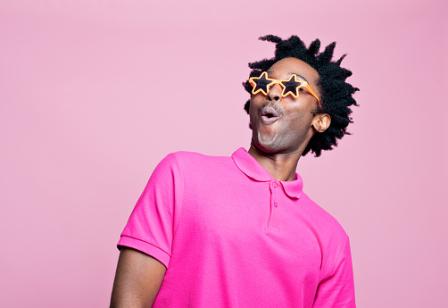 Portrait of excited afro american young man wearing pink polo shirt and star shaped sunglasses, looking away and whistling. Studio shot on pink background.