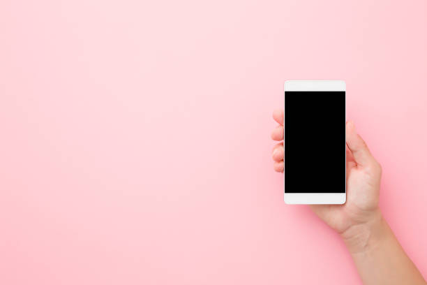 istock Young woman hand holding smartphone with black screen on light pink table background. Pastel color. Empty place for text. Closeup. Top down view. 1209033131