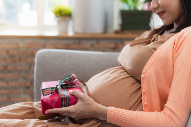 Pregnant hole gift box Pregnant asian woman feeling happy at home while taking care of her child and holding gift box near on her abdomen and smiling while sitting on the couch stomach knot stock pictures, royalty-free photos & images