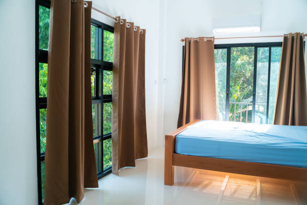 White double bed against window with red curtain. Furniture in bright hostel bedroom. Interior of college dorm.