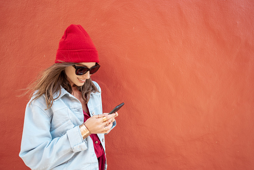 Portrait of a young and cheerful woman stylishly dressed in hat and jacket standing with mobile phone on the red wall background outdoors