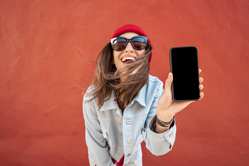 Young stylish woman showing mobile phone while standing relaxed on the red wall background. Phone with black screen to copy paste