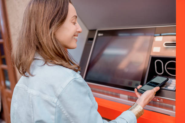 istock Young woman using ATM and mobile phone 1209039585