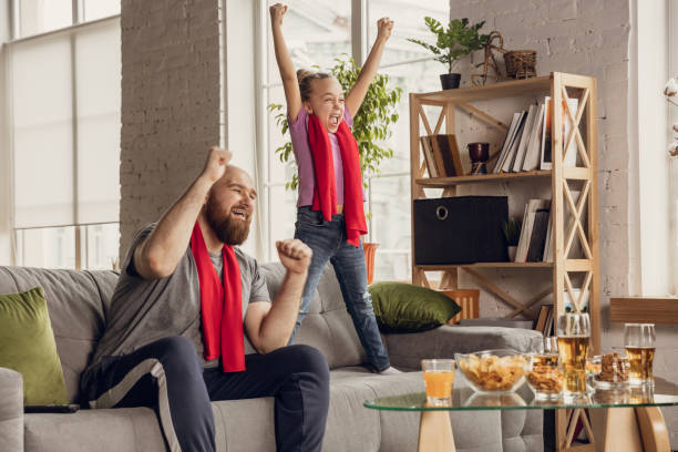 Excited, happy daughter and father watch football, soccer match together on the couch at home Excited, happy daughter and father watching football, soccer, basketball, tennis, hockey match on the couch at home. Fans emotional cheering for favourite national team. Sport, TV, championship. excited football fan cheering for his team on tv stock pictures, royalty-free photos & images
