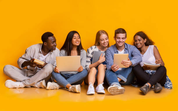 Group of international students studying for university exams Group of multiracial students studying for university exams, sitting on floor, using laptop, digital tablet, holding books, yellow background teenagers only stock pictures, royalty-free photos & images