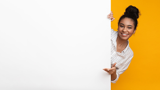 Happy African American Girl Looking Out Behind Blank Advertisement Board With Free Space For Your Text, Yellow Background, Panorama