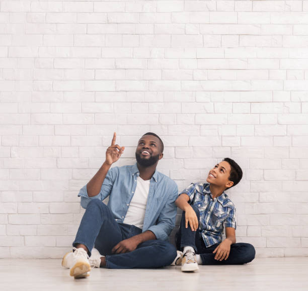 istock Afro Son And Dad Sitting Near Brick Wall And Pointing Upwards 1209053288