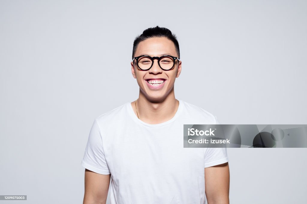 Cheerful asian young man laughing at camera Portrait of happy asian young man wearing white t-shirt and glasses, laughing at camera. Studio shot, white background. Asian and Indian Ethnicities Stock Photo