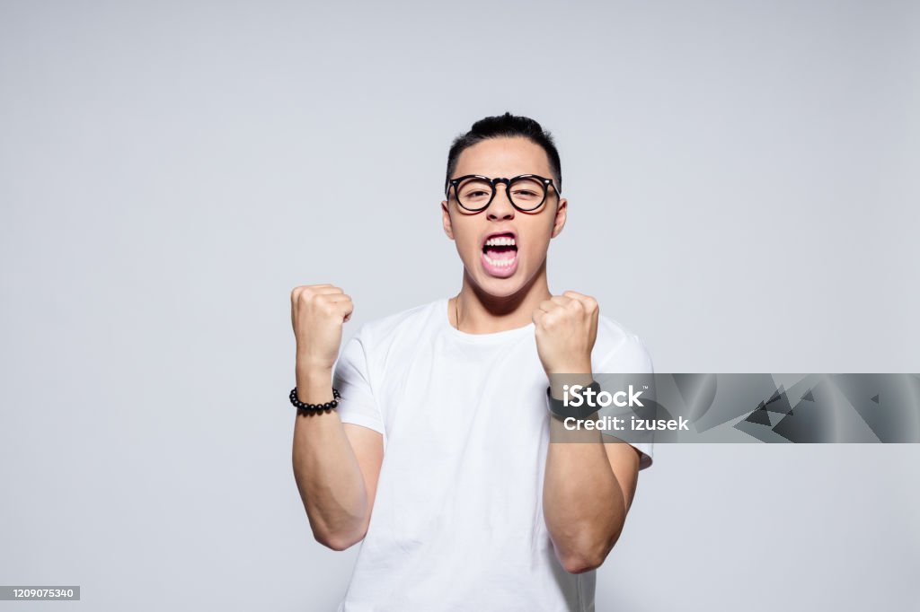 Excited asian winner Portrait of happy asian young man wearing white t-shirt and glasses, in winner gesture. Studio shot, white background. Men Stock Photo