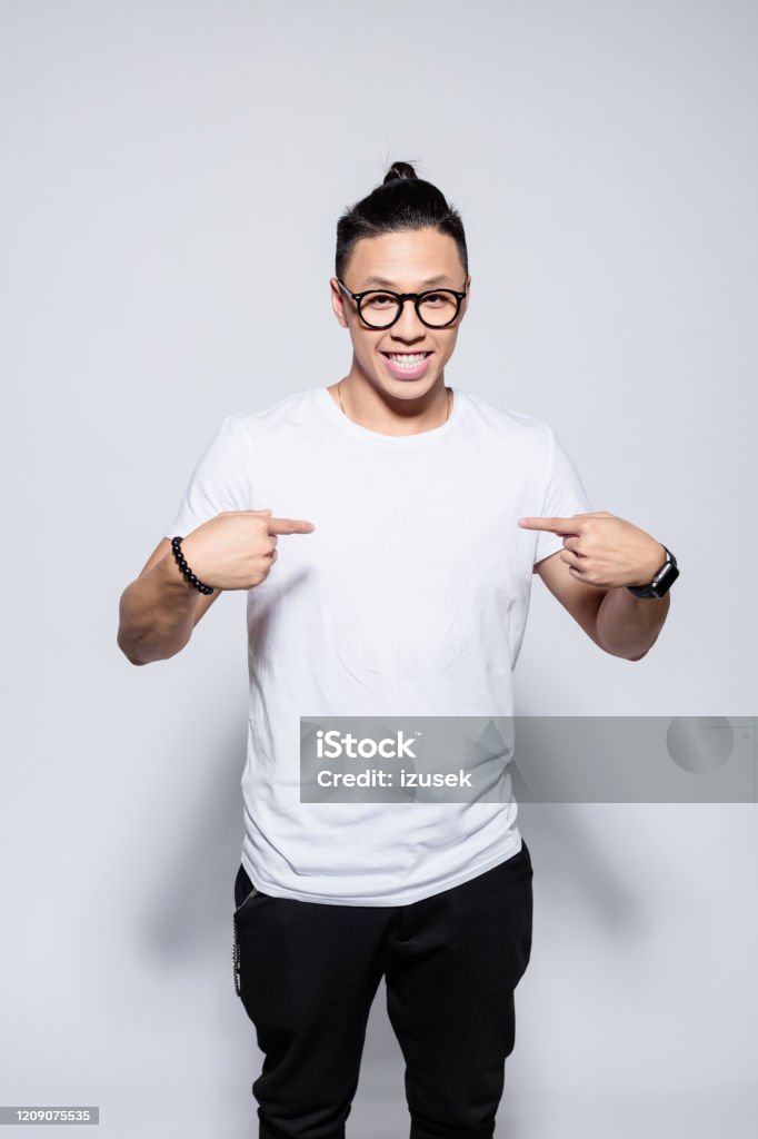 Friendly asian young man pointing at his t-shirt Portrait of happy asian young man wearing white t-shirt and glasses, pointing with index fingers at his chest. Studio shot, white background. White Background Stock Photo