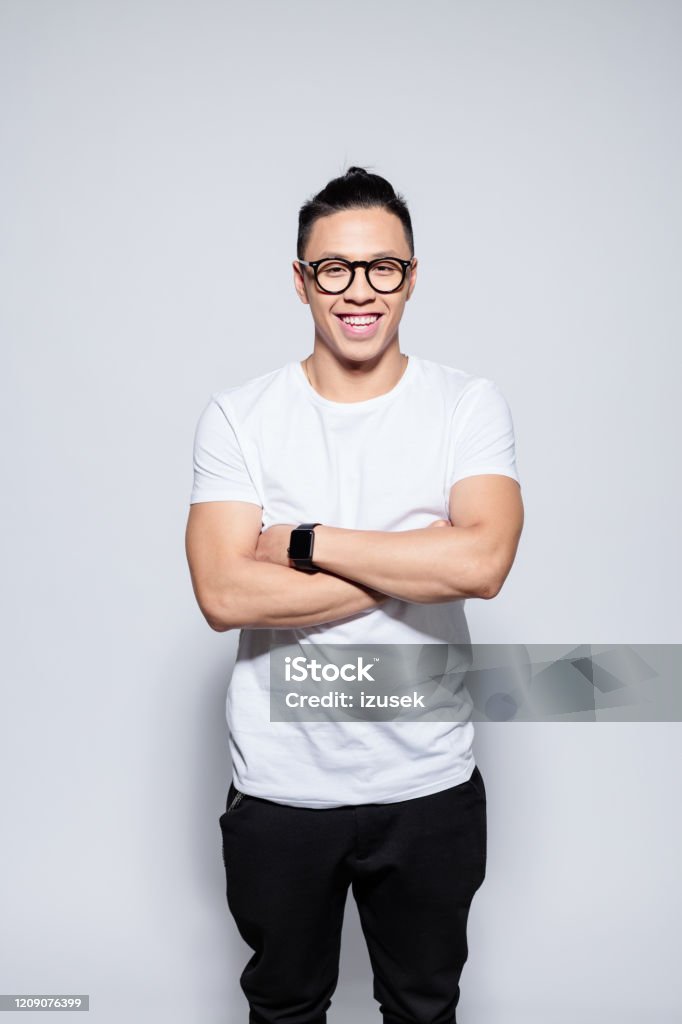 Friendly asian young man standing with arms crossed Portrait of handsome asian young man wearing white t-shirt, black pants and glasses, smiling at camera with arms crossed. Studio shot, white background. Men Stock Photo