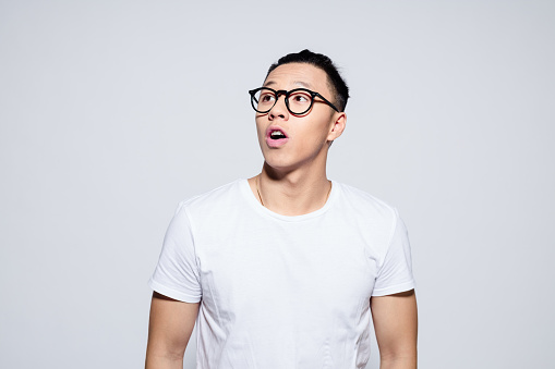 Portrait of surprised asian young man wearing white t-shirt and glasses, looking up with mouth open. Studio shot, white background.