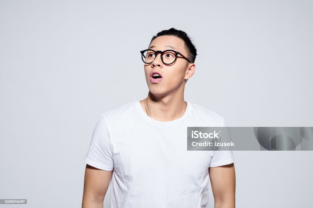 Handsome asian young man looking up Portrait of surprised asian young man wearing white t-shirt and glasses, looking up with mouth open. Studio shot, white background. Men Stock Photo