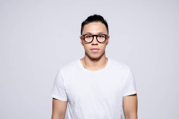 Portrait of serious asian young man wearing white t-shirt and glasses, looking at camera. Studio shot, white background.