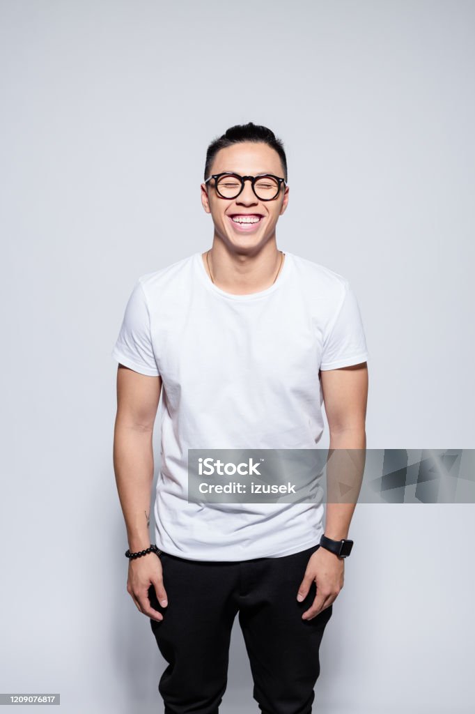 Happy asian young man laughing at the camera Portrait of handsome asian young man wearing white t-shirt, black pants and glasses, laughing at camera. Studio shot, white background. Men Stock Photo