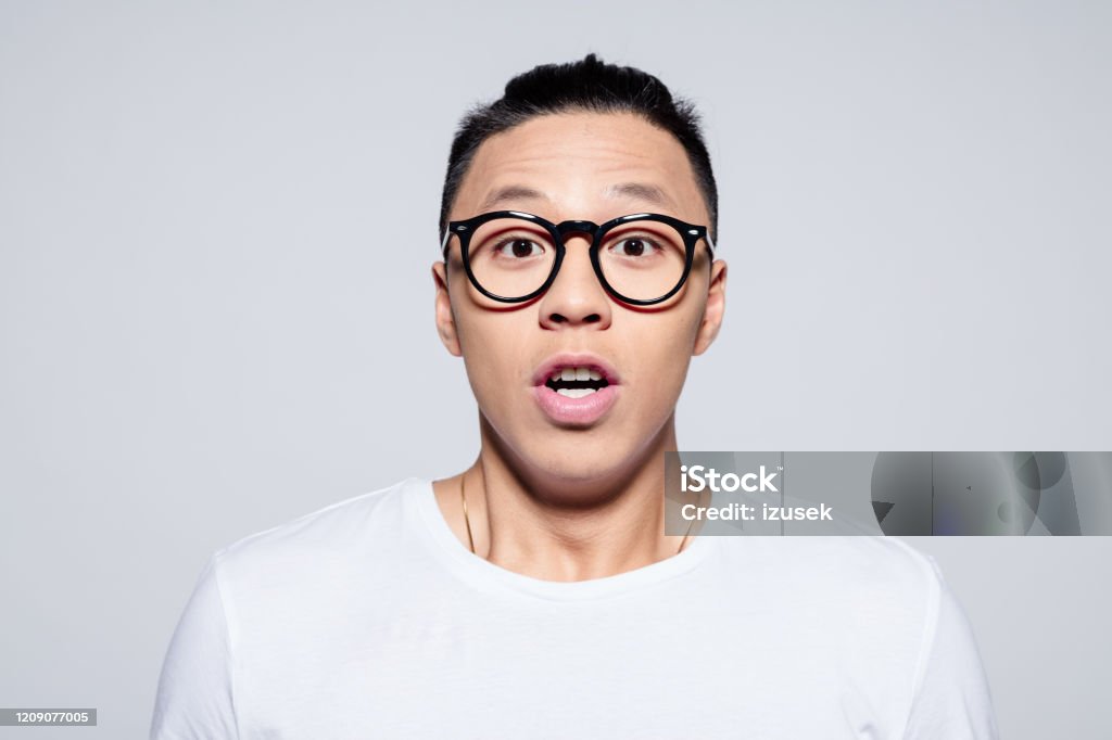Headshot of surprised asian young man Portrait of handsome asian young man wearing white t-shirt and glasses, gasping at camera, rolling eyes. Studio shot, white background. Looking At Camera Stock Photo