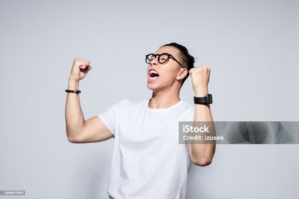Portrait of excited asian winner Portrait of happy asian young man wearing white t-shirt and glasses, in winner gesture. Studio shot, white background. Asian and Indian Ethnicities Stock Photo