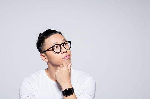 Headshot of worried asian young man wearing white t-shirt and glasses, looking at copy space with hand on chin. Studio portrait on white background.