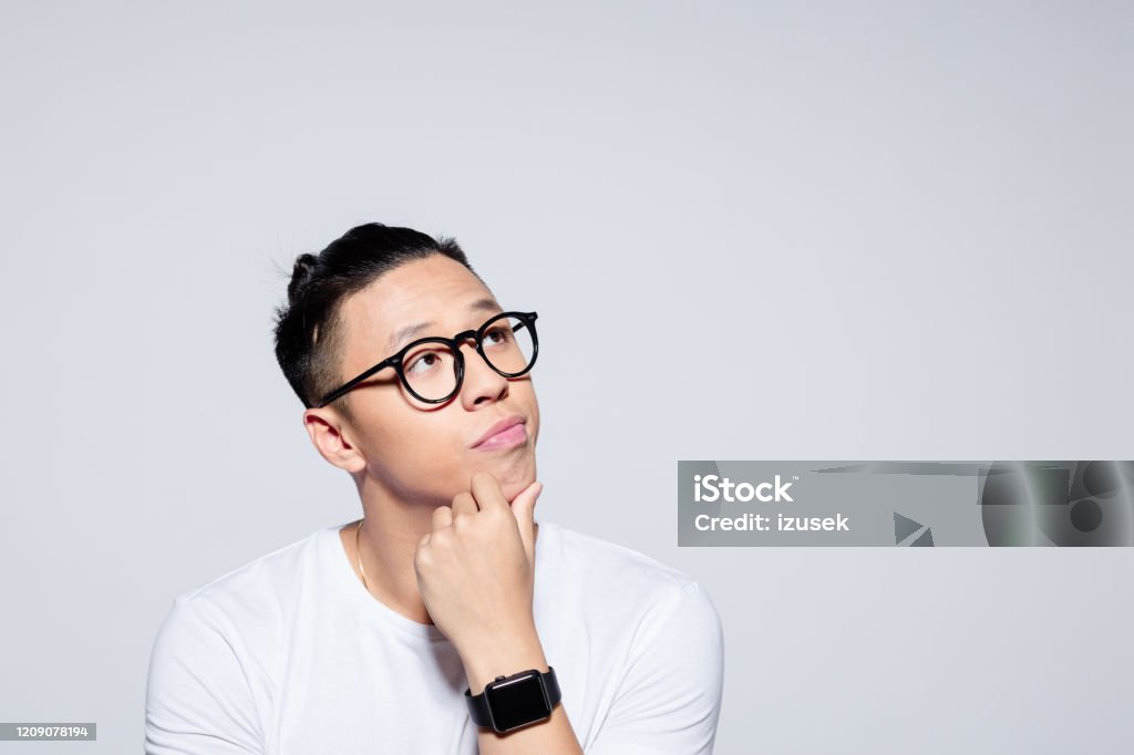 Portrait of pensive asian young man Headshot of worried asian young man wearing white t-shirt and glasses, looking at copy space with hand on chin. Studio portrait on white background. Contemplation Stock Photo