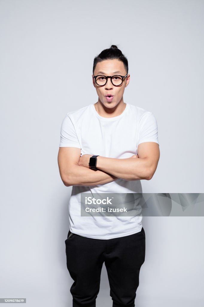 Surprised asian young man standing with arms crossed Portrait of handsome asian young man wearing white t-shirt, black pants and glasses, looking at camera with arms crossed. Studio shot, white background. Men Stock Photo