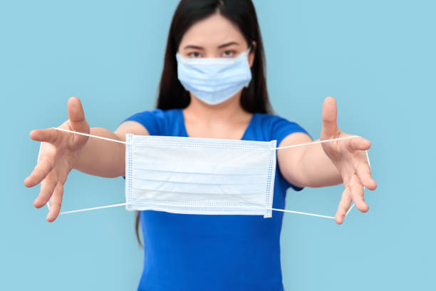 Coronavirus Concept. Chinese woman in medical mask standing isolated on grey givinganother mask to camera close-up stock photo