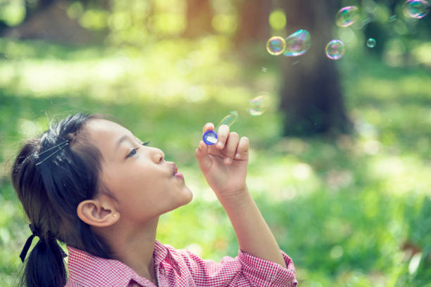 夏の屋外で緑の遊び場で愛らしい幸せなかわいいかわいい女の子の遊び心泡。公園の幸福の時間で面白い陽気な女の子。自然公園で遊び心のある石鹸を吹く泡。 - bubble wand child blowing asian ethnicity ストックフォトと画像