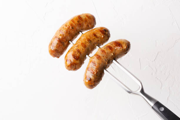 istock Fried meat sausages with spices and herbs on big fork on white background 1209100230