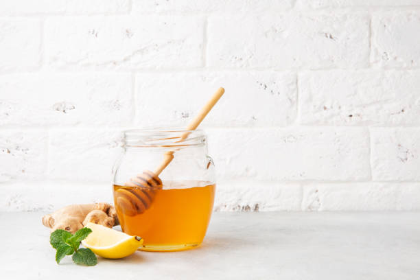 miel en un frasco de vidrio con cuchara de madera y panal, limón, jengibre y menta sobre una mesa gris y fondo de pared de ladrillo blanco - fruit winter orange lemon fotografías e imágenes de stock