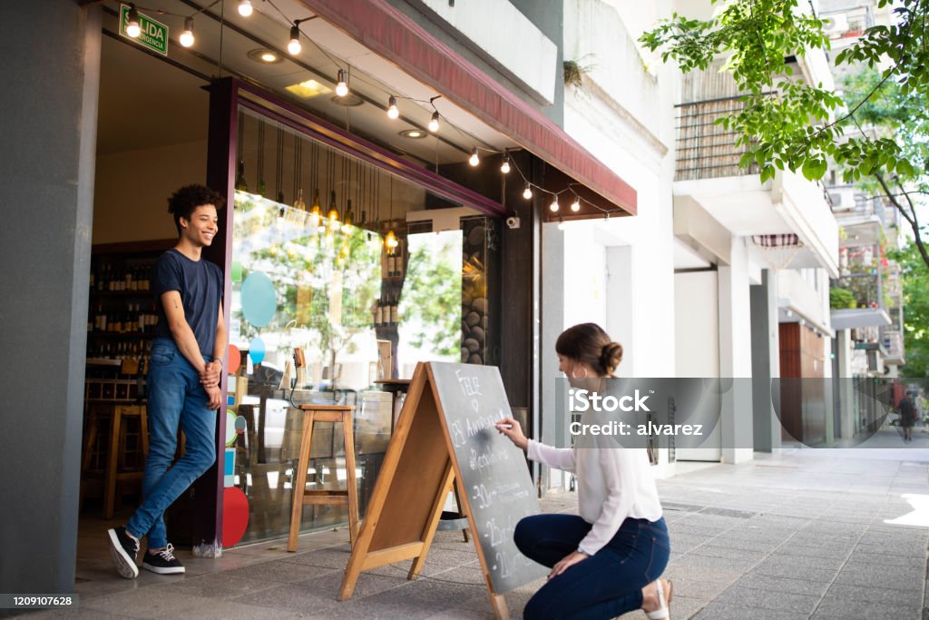 Employees working at a winery store Woman writing on a chalkboard outdoors with man standing at door of wine shop. Employees working at a winery store. Store Stock Photo