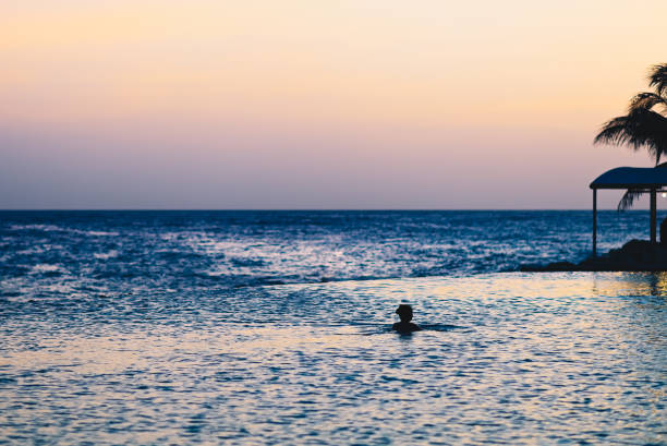 A lone person in silhouette enjoys an infinity pool at dusk that seems to blend directly in with the cast ocean beyond. Stunning sunset over the tropical island of Curacao, tranquility, relaxation and the beauty of nature align for an idyllic moment