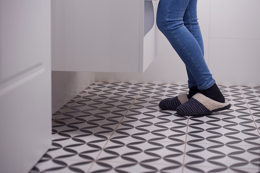 Low section of a female`s feet wearing jeans, black socks, and warm slipper, standing in front of the sink cabinet in the bathroom.