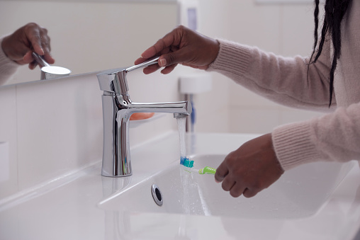 Close up image of a woman's hands opening the faucet to wash teeth-brush in the white ceramic sink under the running water in the bathroom.