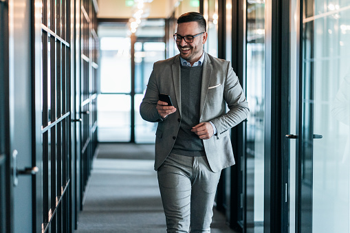 Young smiling businessman using smart phone while walking in the office corridor