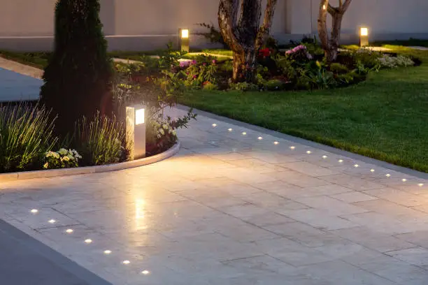 Photo of marble tile playground in the night backyard of mansion with flowerbeds and lawn with ground lamp and lighting in the warm light at dusk in the evening.