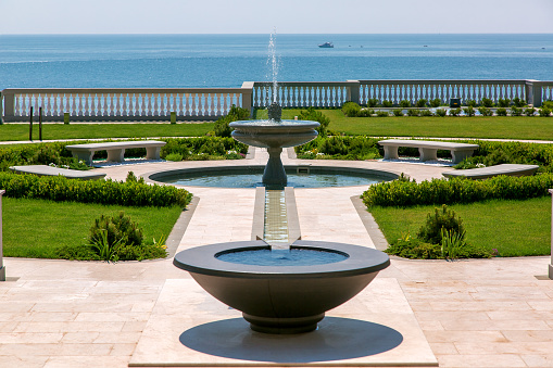the backyard of a landscaped mansion with a stone fountain and stream along a marble path with a green lawn and plants, in the background is the blue sea on a sunny summer day.