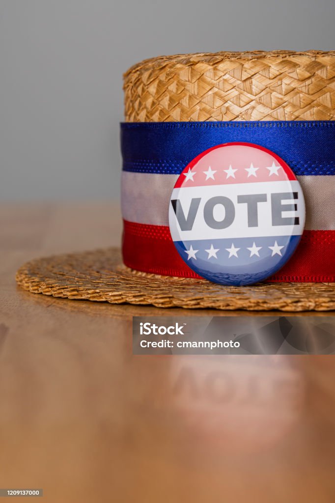 A skimmer hat with a red, white and blue head band and a VOTE button sitting on a wood table A low angle view of a straw Skimmer hat with a red, white and blue head band and a VOTE button on the front sitting on a wood table in a precinct. American Flag Stock Photo