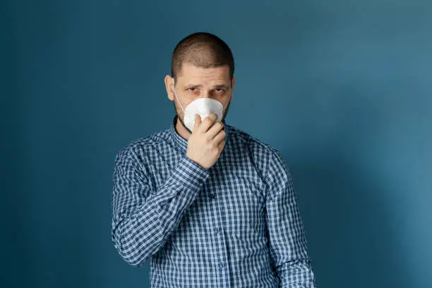 Portrait of adult caucasian man standing in front of the blue wall wearing shirt and holding protective mask on his face against virus flue bacteria protection health issues front view