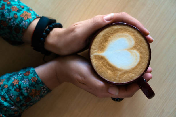 female hands hugging a large mug with cappuccino. heart-shaped latte art - espresso women cup drink imagens e fotografias de stock