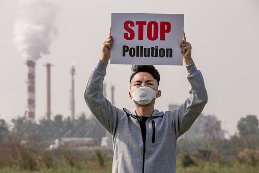 asian man hold stop pollution sign and wears protective n95 mask against air pollution standing in front of factory