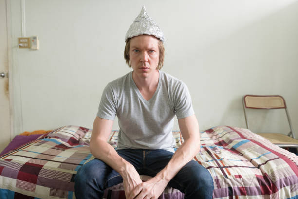 Portrait of young handsome man wearing tin foil hat as conspiracy theory concept in the bedroom at home